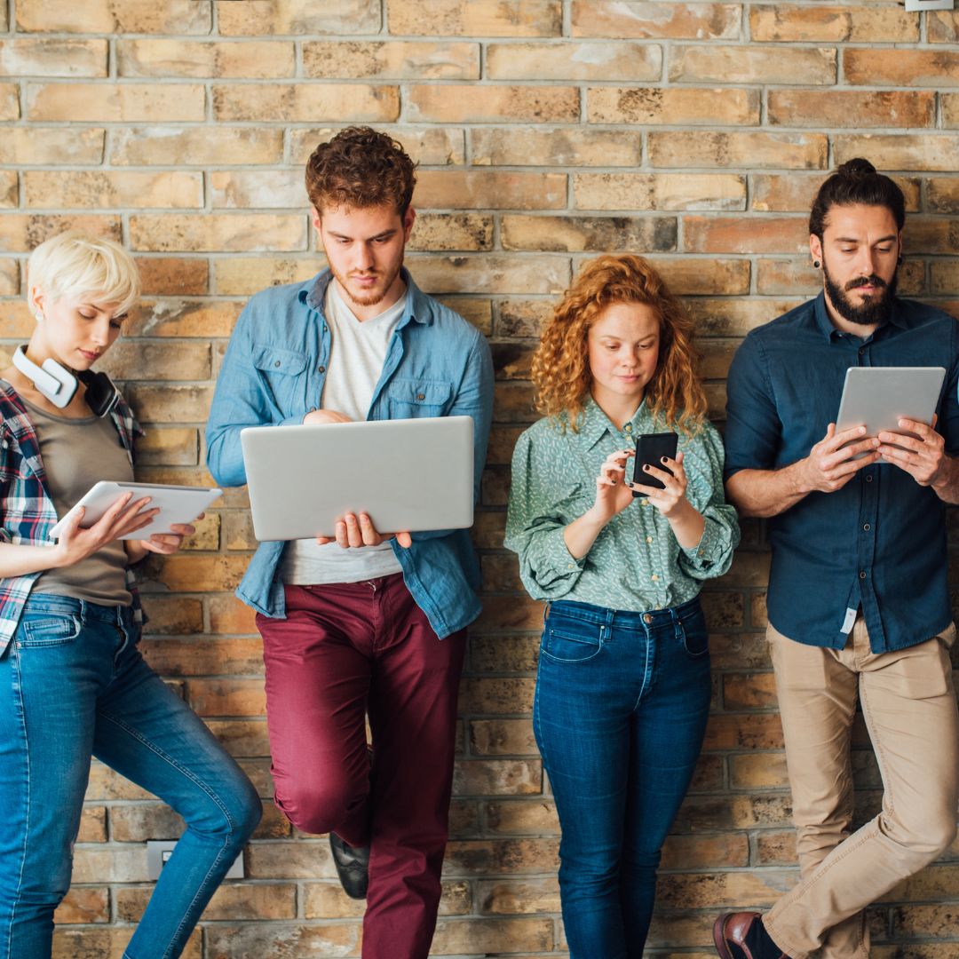 Group of millennials holding digital devices while standing against a wall